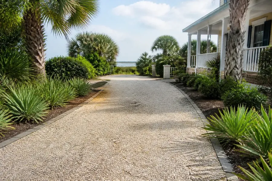 Light-colored crushed-shell driveway bordered by pavers, palms, and low green plants beside a house with a white porch, with water visible in the distance.