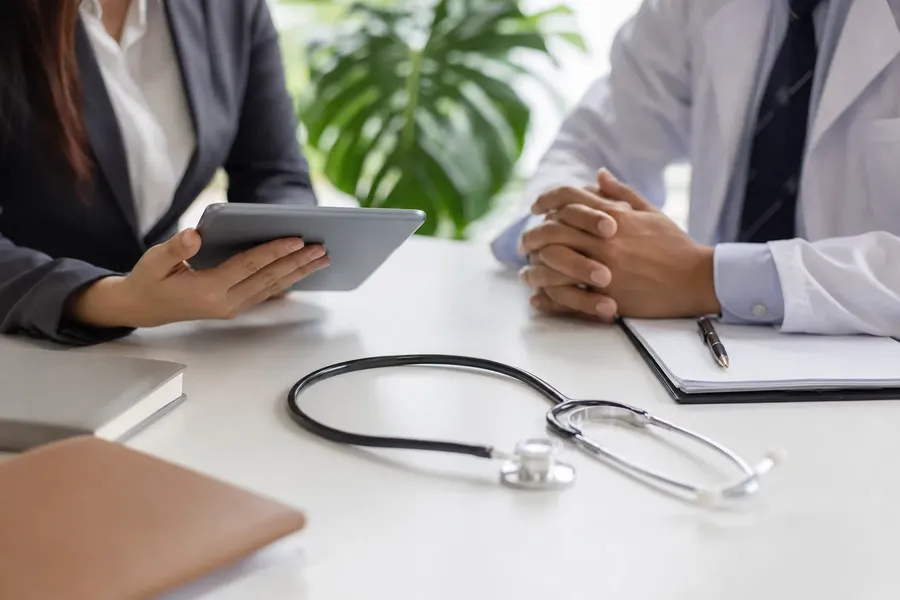 Clinician and Patient Meeting at a Desk A clinician and a patient sit across a desk with a clipboard and stethoscope between them.