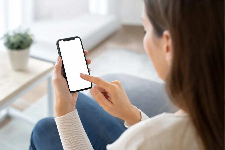Woman Touching a Smartphone With a Blank Screen A seated woman holds a smartphone with a blank white screen and touches it with her index finger, viewed over her shoulder.